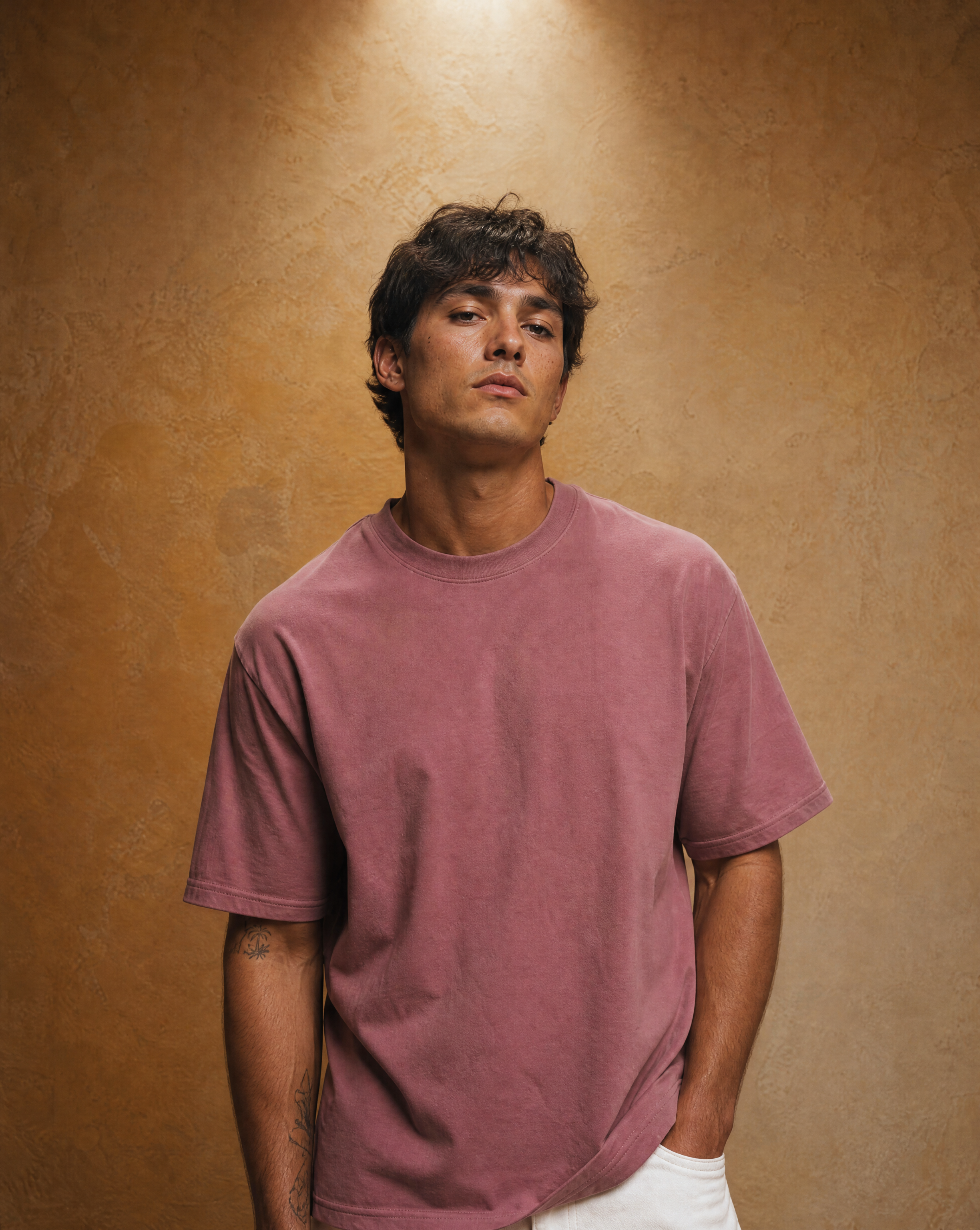 A man wearing a washed, muted dusty rose short-sleeve t-shirt and white pants, standing against a textured warm brown studio backdrop.