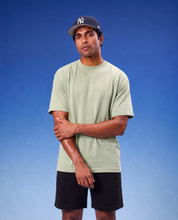 A man wearing a light sage green crewneck t-shirt, black shorts, and a navy blue New York Yankees baseball cap, standing against a blue gradient background.