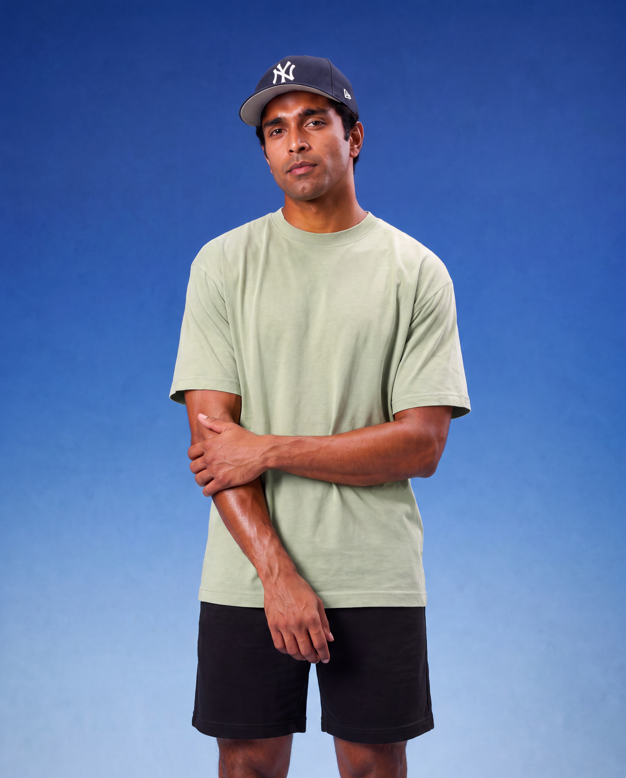 A man wearing a light sage green crewneck t-shirt, black shorts, and a navy blue New York Yankees baseball cap, standing against a blue gradient background.