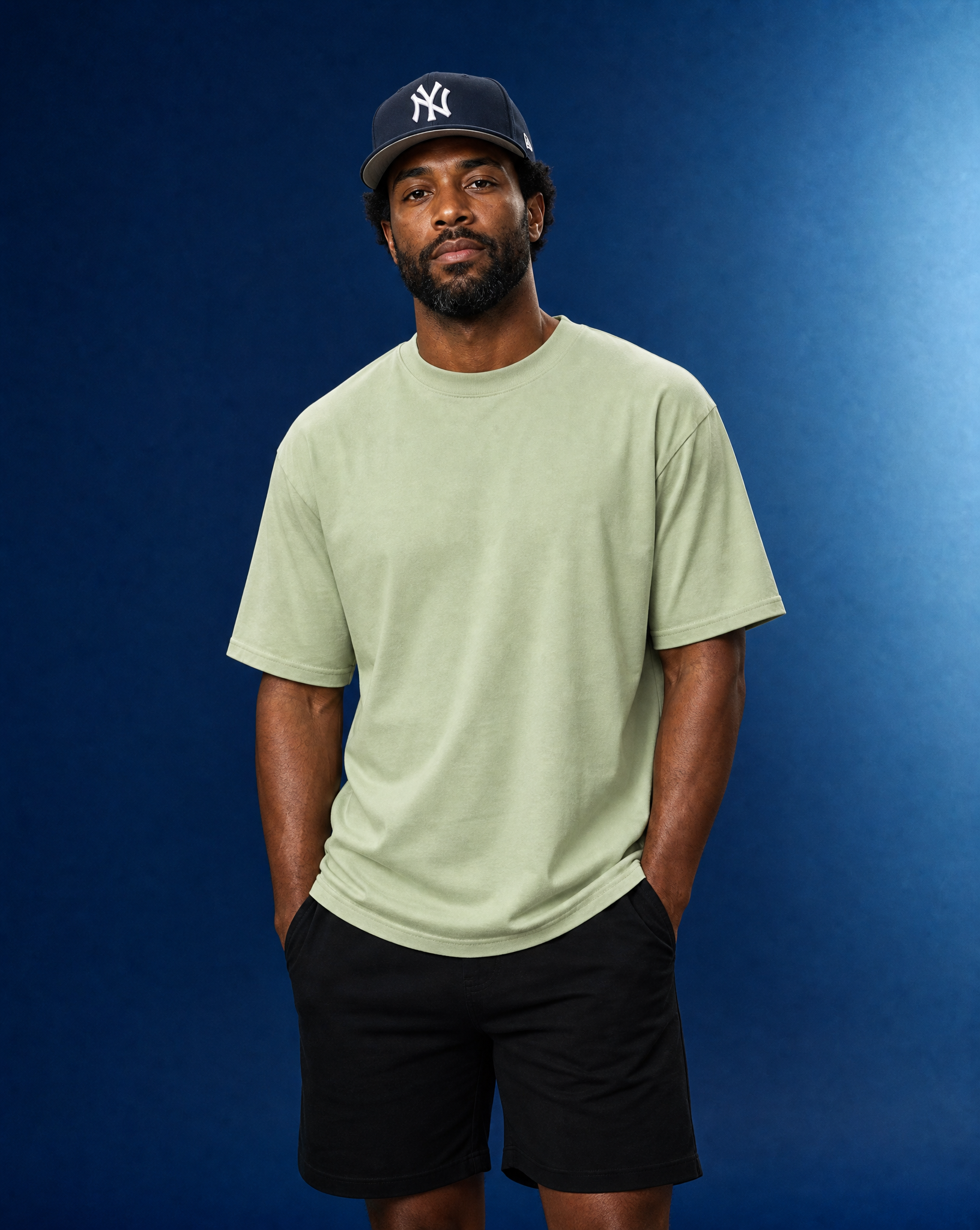 A man wearing a light sage green crewneck t-shirt, black shorts, and a navy blue New York Yankees baseball cap, standing against a dark blue studio background.