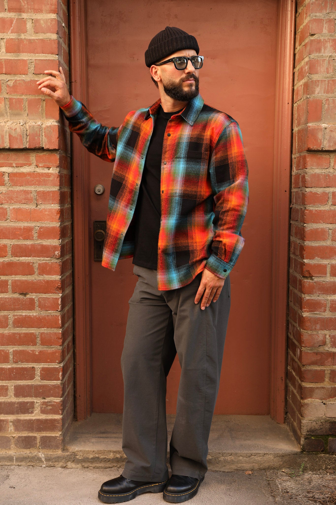 A man wearing a vibrant orange, red, and blue plaid button-down shirt over a black t-shirt, gray pants, and a black beanie stands in front of a rustic red-brown door framed by brick walls.