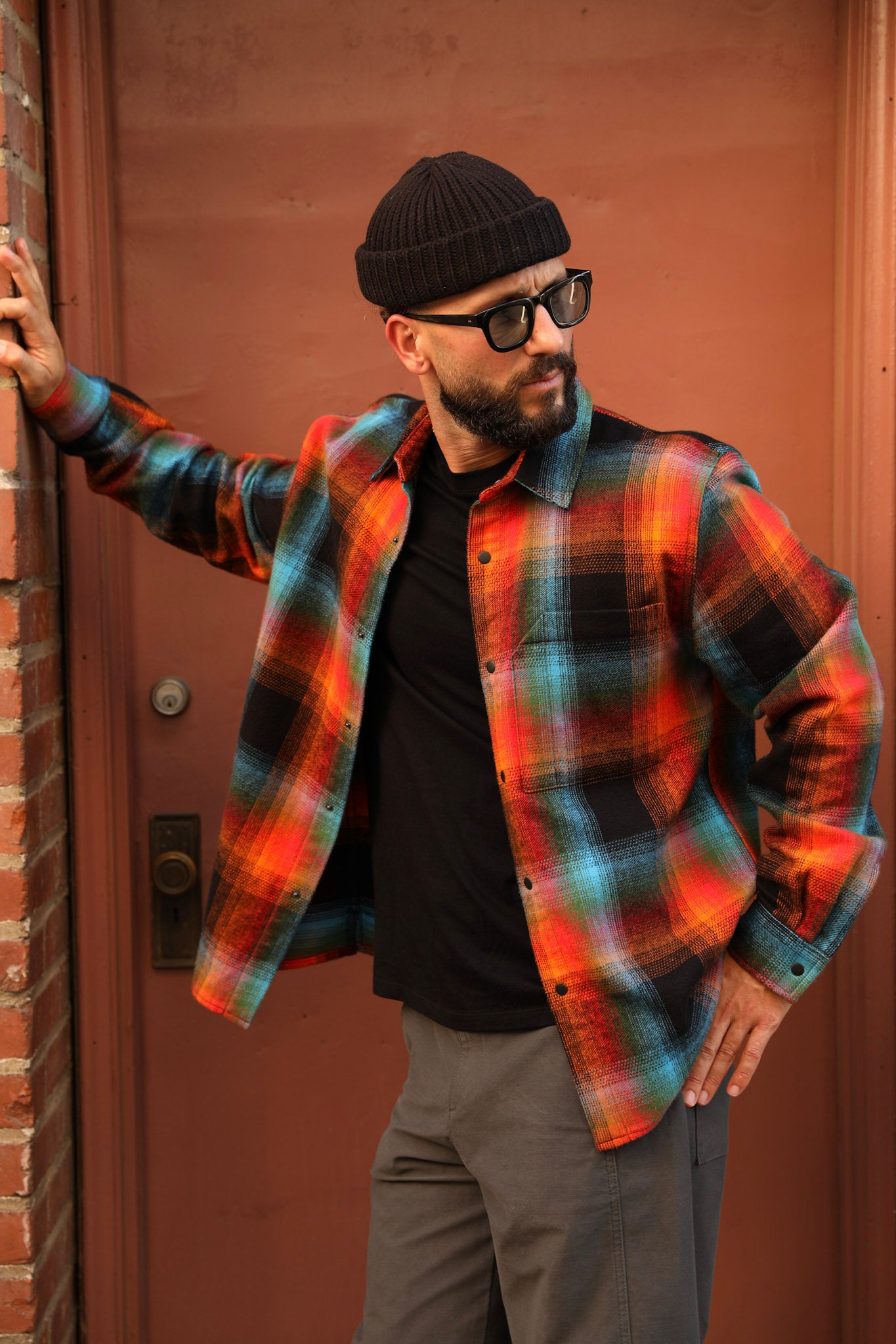 A man wearing a vibrant orange, red, and blue plaid button-down shirt over a black t-shirt, gray pants, and a black beanie, posing in front of a rustic reddish-brown wooden door.