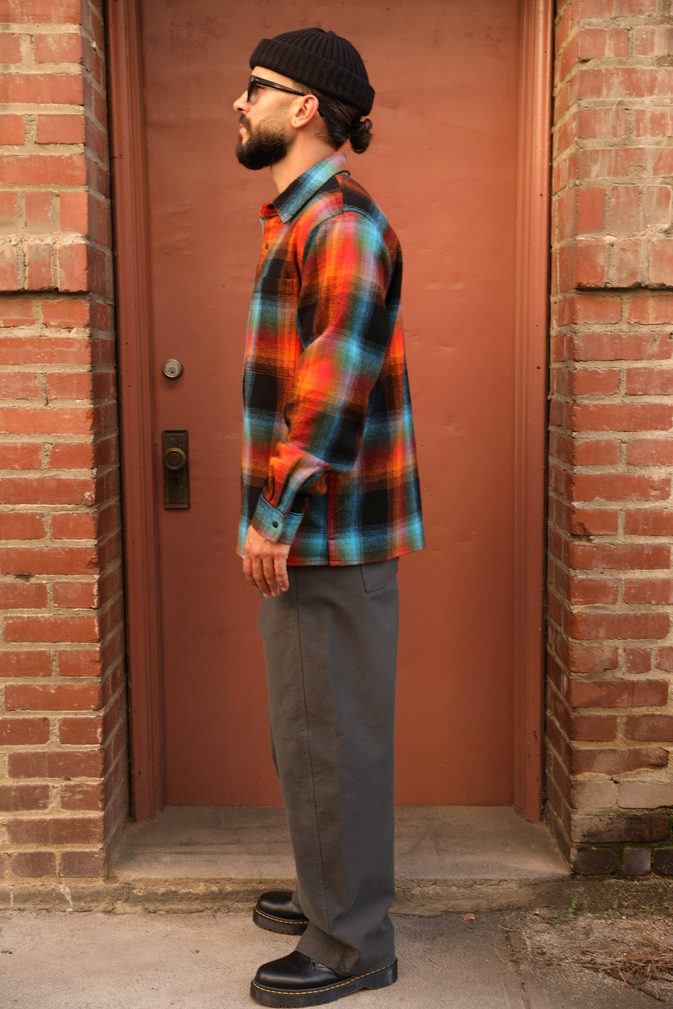 A man wearing a colorful orange, blue, and green plaid flannel shirt, gray pants, and a black beanie stands in profile in front of a reddish-brown wooden door framed by brick walls.