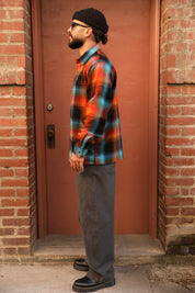 A man wearing a colorful orange, blue, and green plaid flannel shirt, gray pants, and a black beanie stands in profile in front of a reddish-brown wooden door framed by brick walls.
