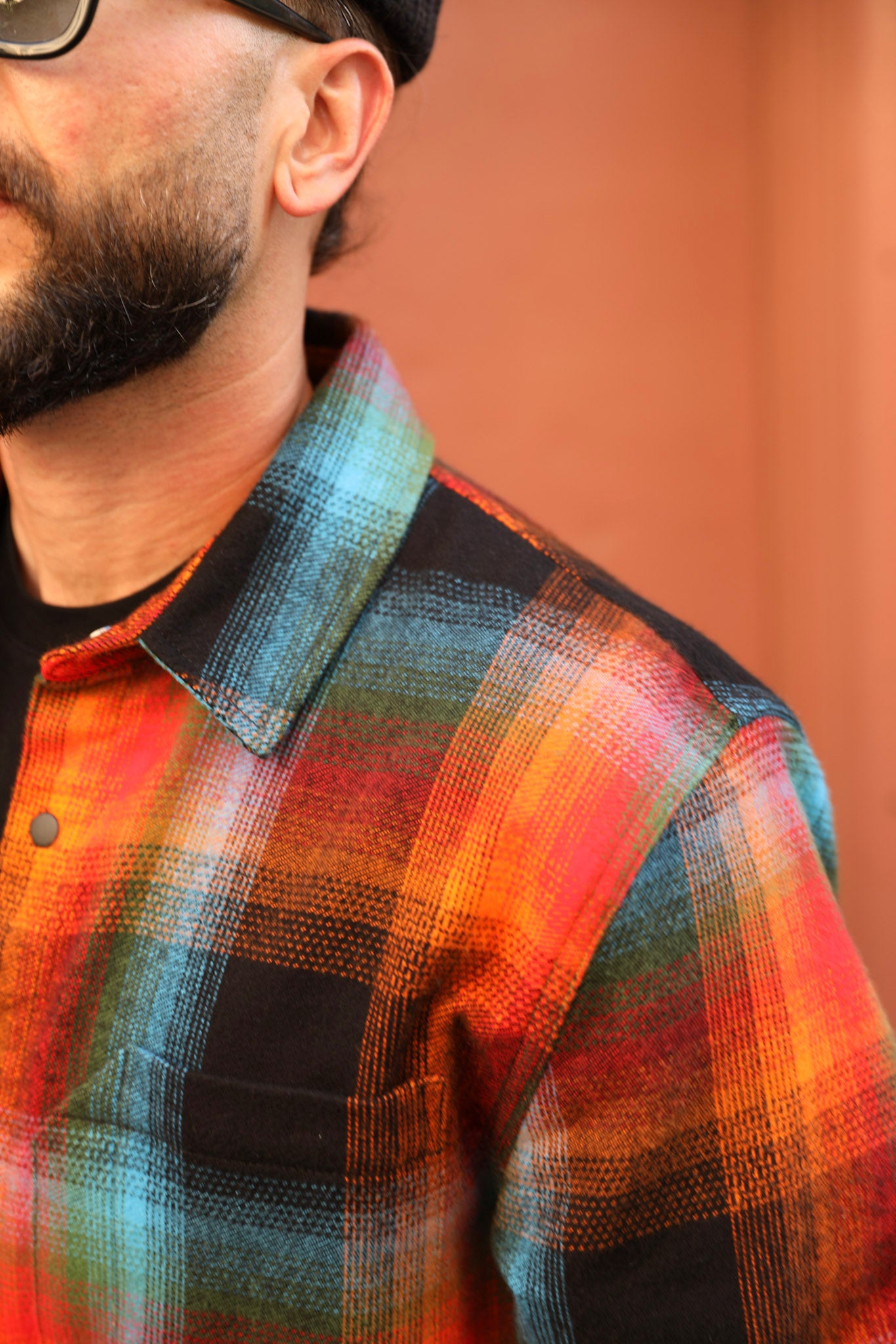 Close-up of a man wearing a colorful plaid flannel shirt with orange, red, blue, and black checkered patterns, set against a warm terracotta background.