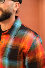 Close-up of a man wearing a colorful plaid flannel shirt with orange, red, blue, and black checkered patterns, set against a warm terracotta background.