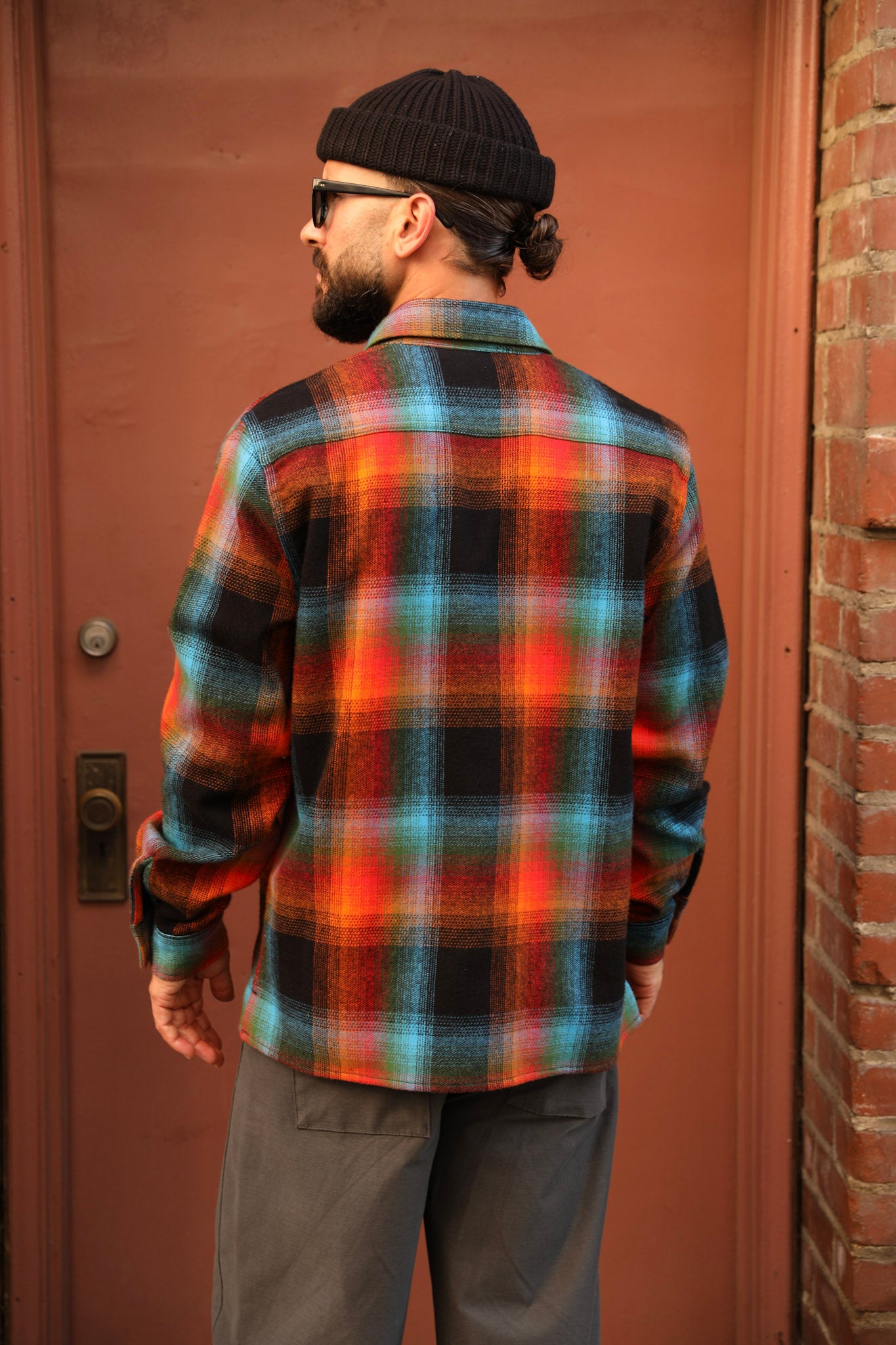 A man wearing a vibrant multicolor plaid flannel shirt, black beanie, and sunglasses, standing in front of a rustic reddish-brown wooden door.