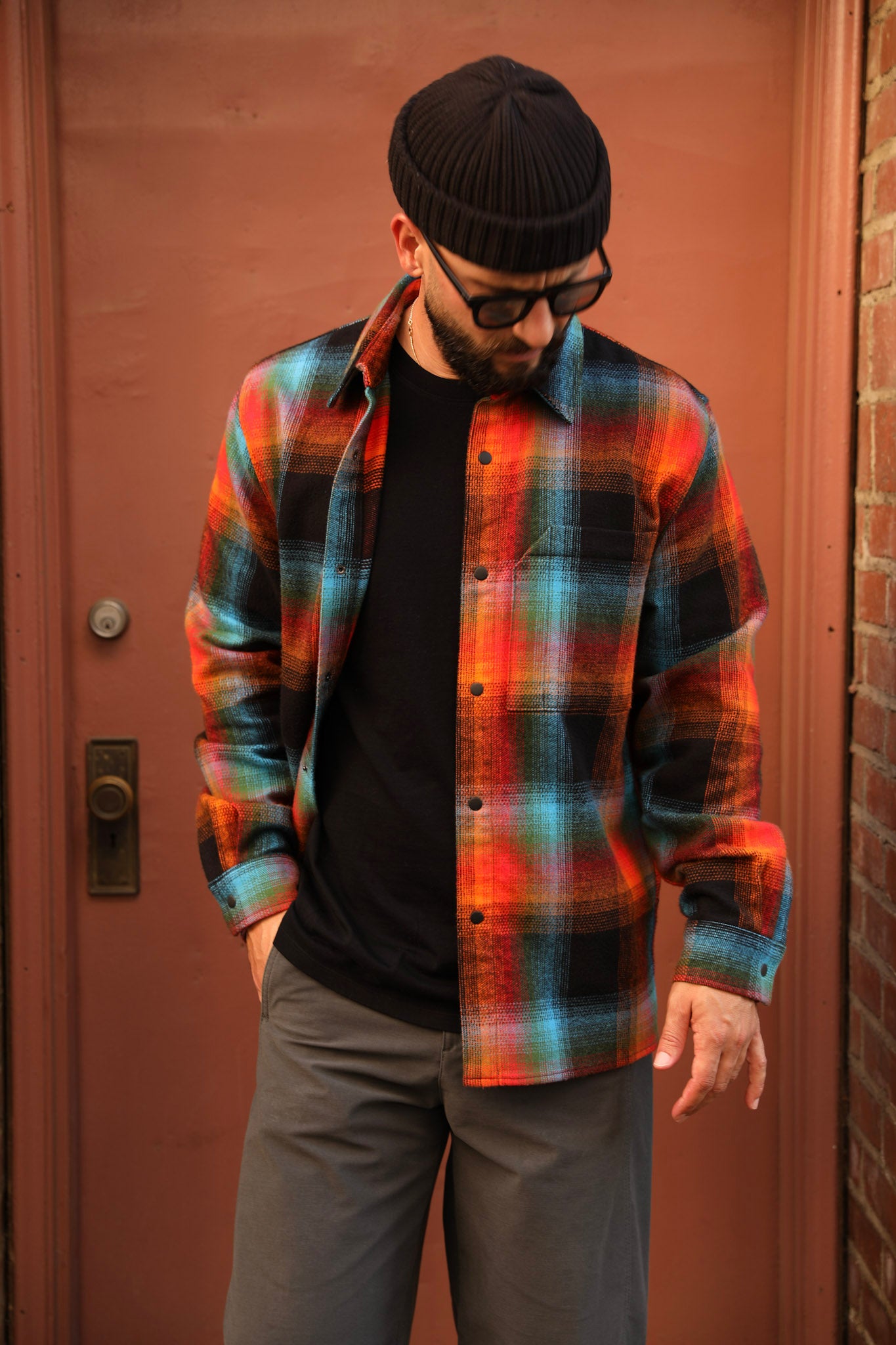 A man wearing a vibrant orange, red, and blue plaid flannel shirt over a black t-shirt and gray pants, standing in front of a reddish-brown door.