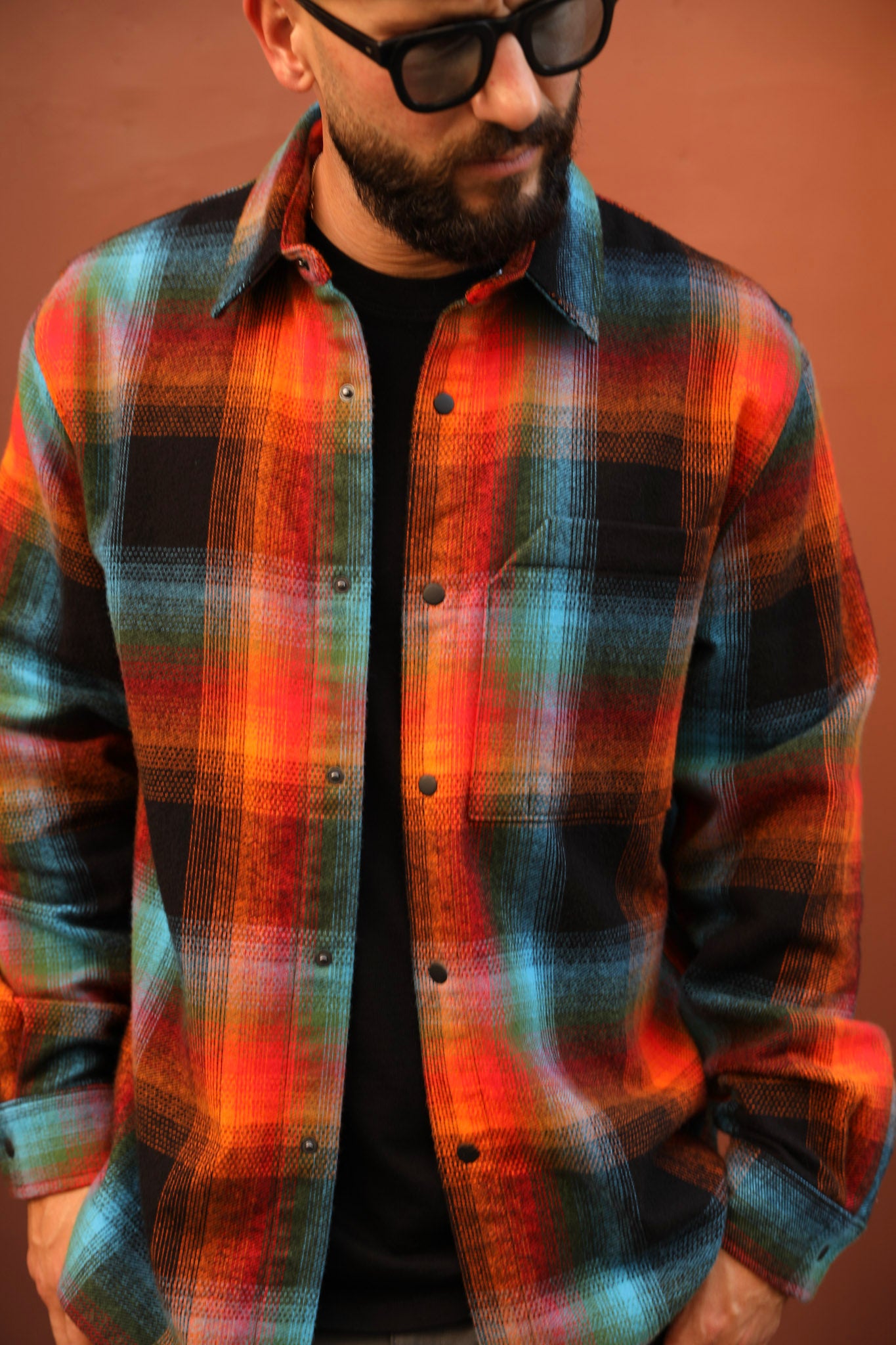 A man wearing a vibrant orange, red, and teal plaid button-up flannel shirt over a black t-shirt, with black sunglasses, set against a solid terracotta background.