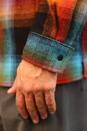 Close-up of a person's hand wearing a multicolor plaid flannel shirt with a blue cuff and a black snap button, paired with dark gray pants.