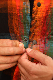 Close-up of a person's hands buttoning a colorful plaid flannel shirt with orange, red, green, and black stripes.