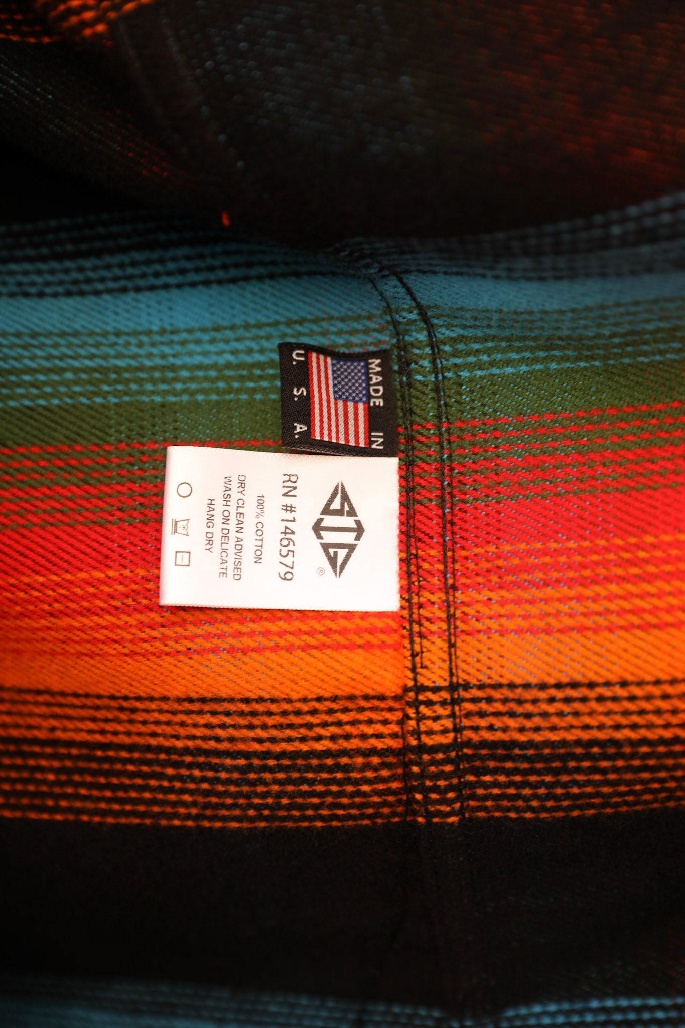 Close-up of a plaid garment label sewn onto a striped textile with black, teal, green, orange, and red tones.