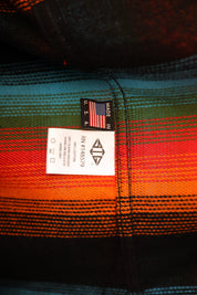 Close-up of a plaid garment label sewn onto a striped textile with black, teal, green, orange, and red tones.