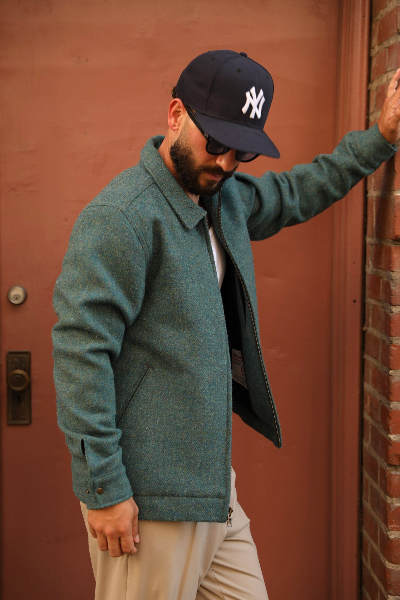 A man wearing a textured sage green wool-blend jacket, beige pants, a black New York Yankees baseball cap, and sunglasses, posing against a reddish-brown door and brick wall.
