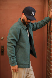 A man wearing a textured sage green wool-blend jacket, beige pants, a black New York Yankees baseball cap, and sunglasses, posing against a reddish-brown door and brick wall.