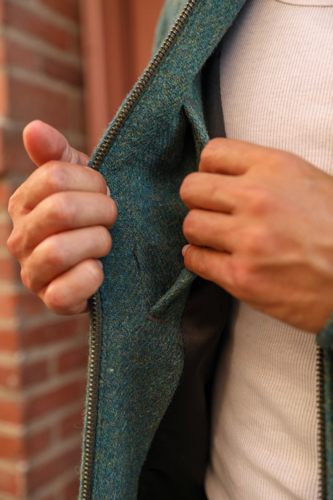 Close-up of a person wearing a textured teal-green wool-blend zip jacket over a cream ribbed sweater, with hands opening the jacket to show the lining.