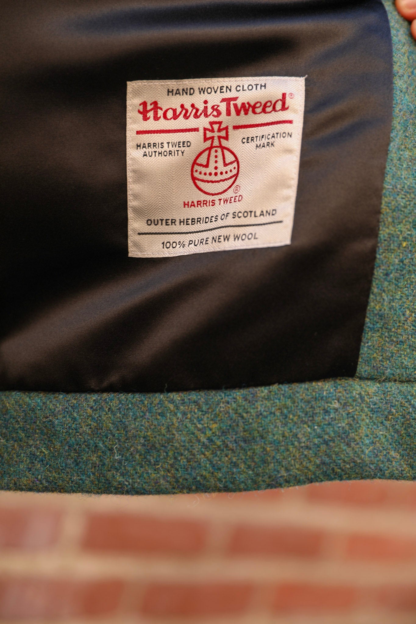 Close-up of a fabric label reading Harris Tweed on a dark satin-like backing, with a blue-green woven textile surface below and a blurred brick wall in the foreground.