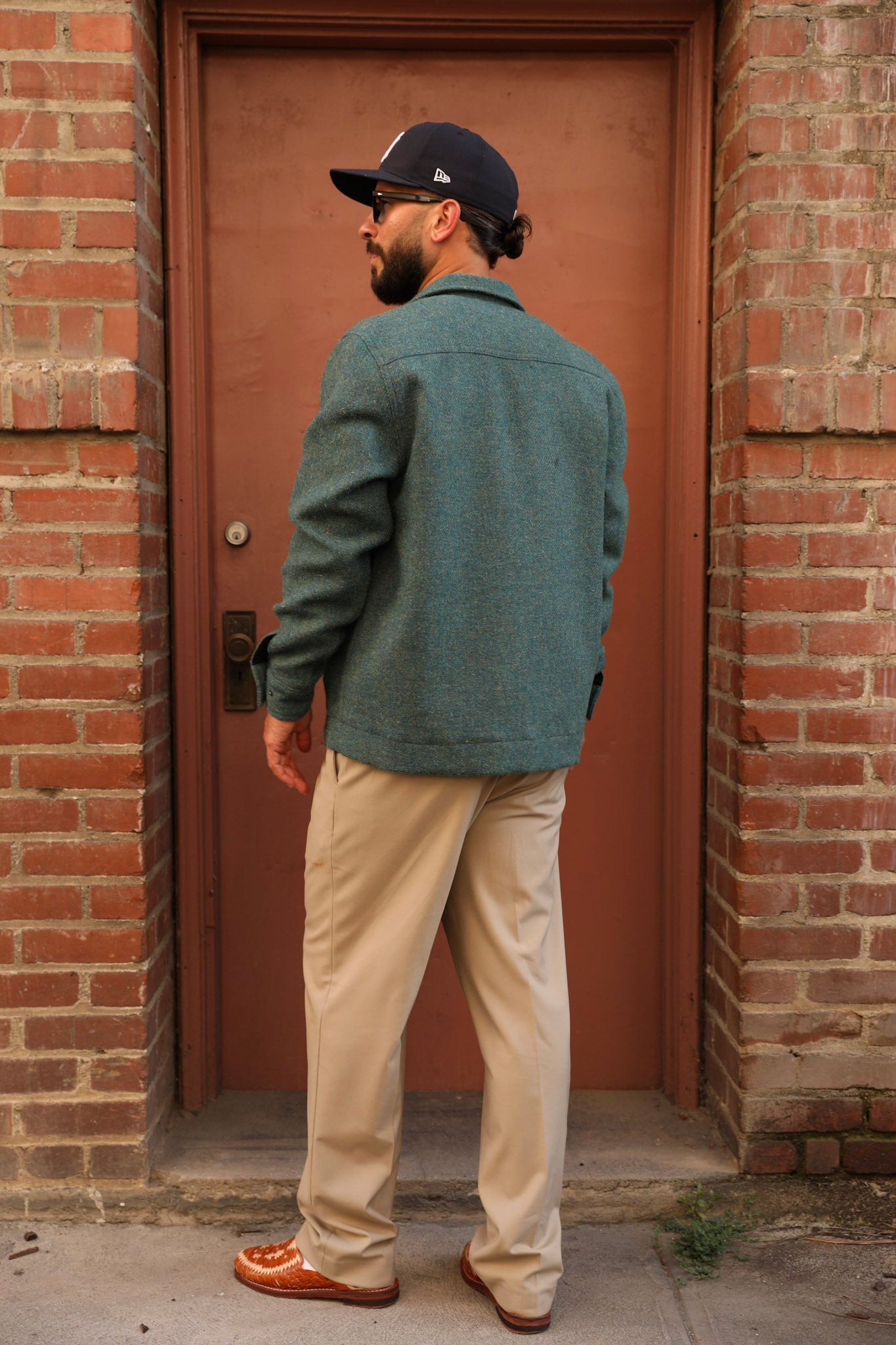 A man wearing a teal green textured overshirt, beige trousers, and a black baseball cap stands in profile in front of a rustic red-brown door framed by red brick walls.