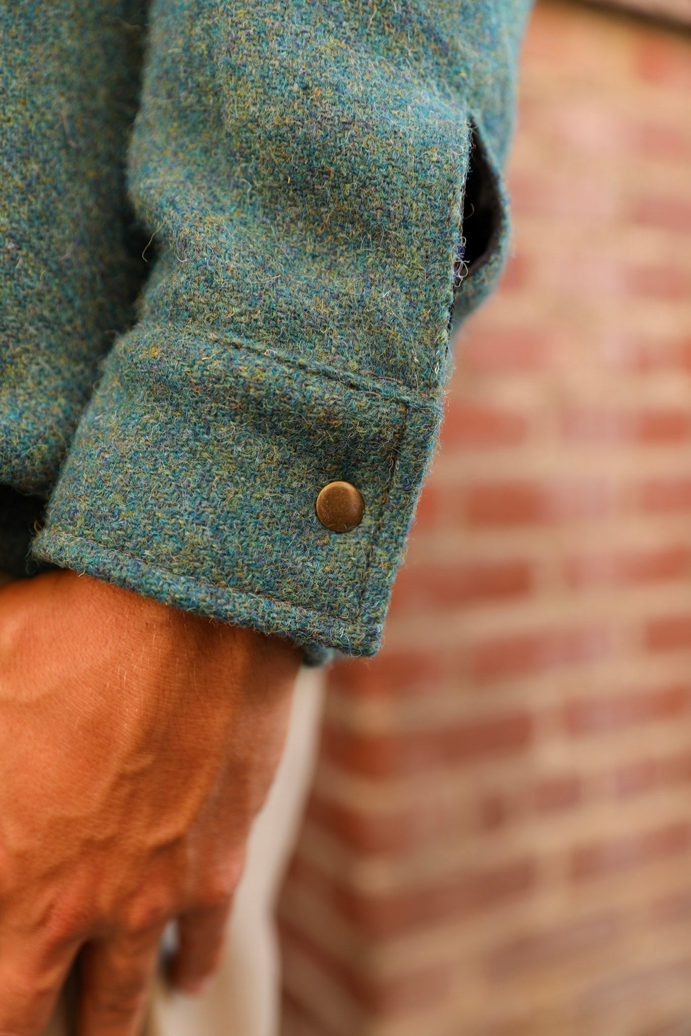 Close-up of a person wearing a textured teal tweed jacket, showing the sleeve with a button cuff and a hand, with a blurred brick wall in the background.