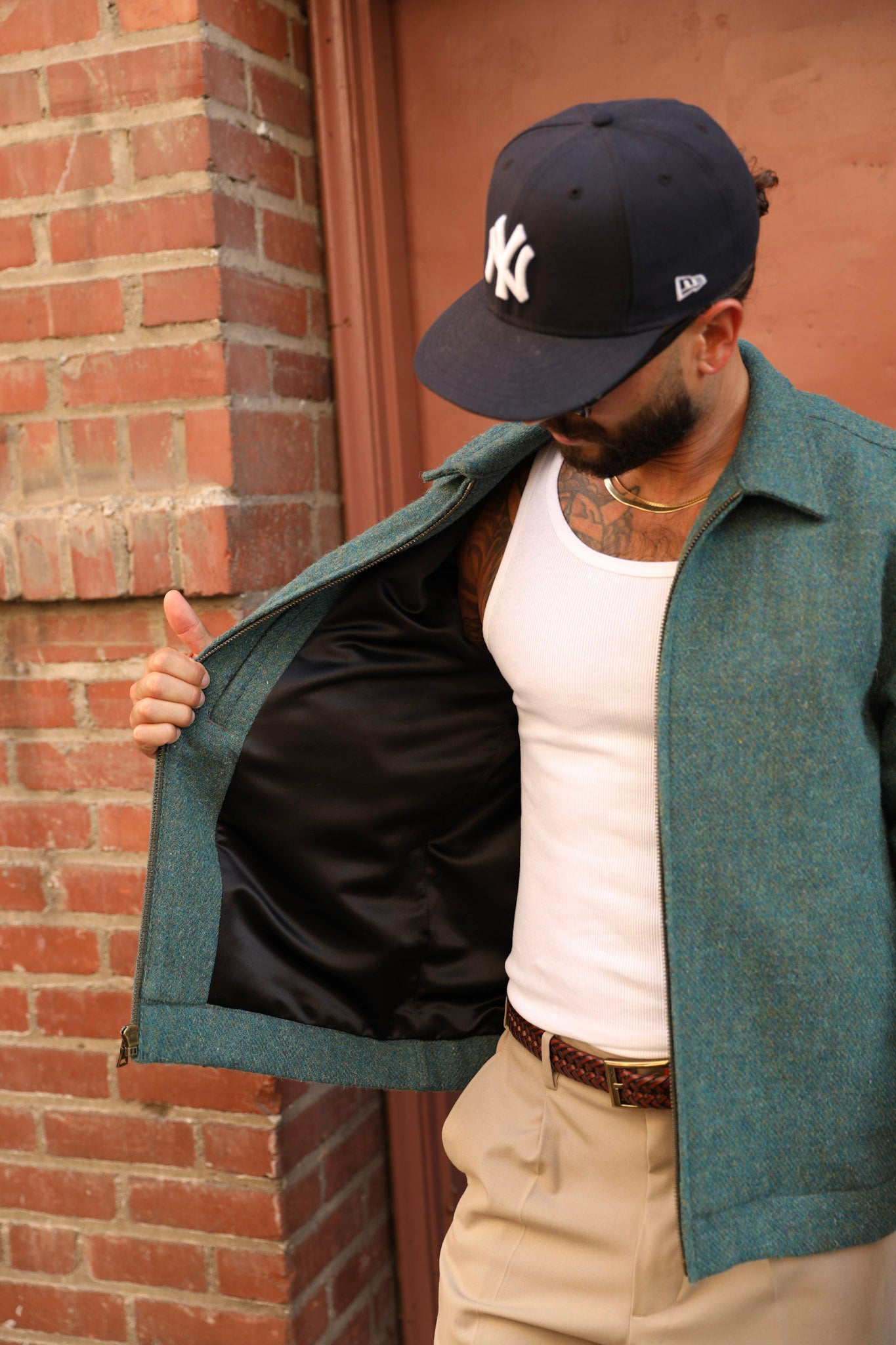 A man wearing a teal green textured jacket with a black satin lining, a white tank top, khaki pants, and a black New York Yankees baseball cap, standing in front of a brick wall.