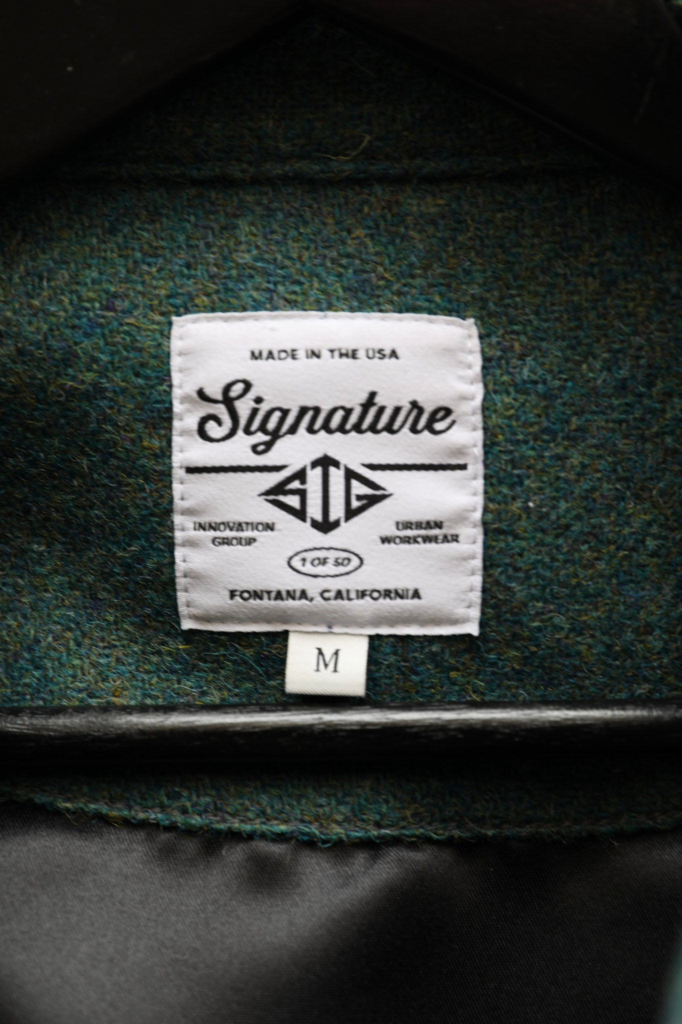 Close-up of a woven wool label reading 'Signature' and 'Made in the USA' sewn onto the inside collar of a dark green textured jacket, with a small size tag below.
