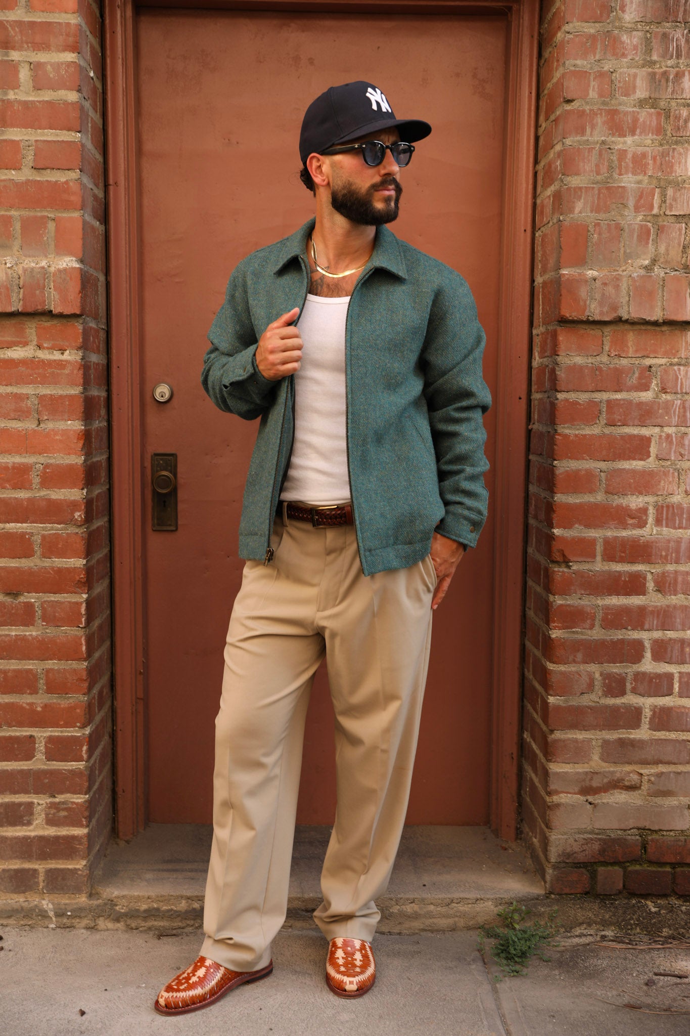 A man wearing a dark green overshirt, beige trousers, a white tank top, a black baseball cap, sunglasses, and brown loafers stands in front of a brick building with a brown door.