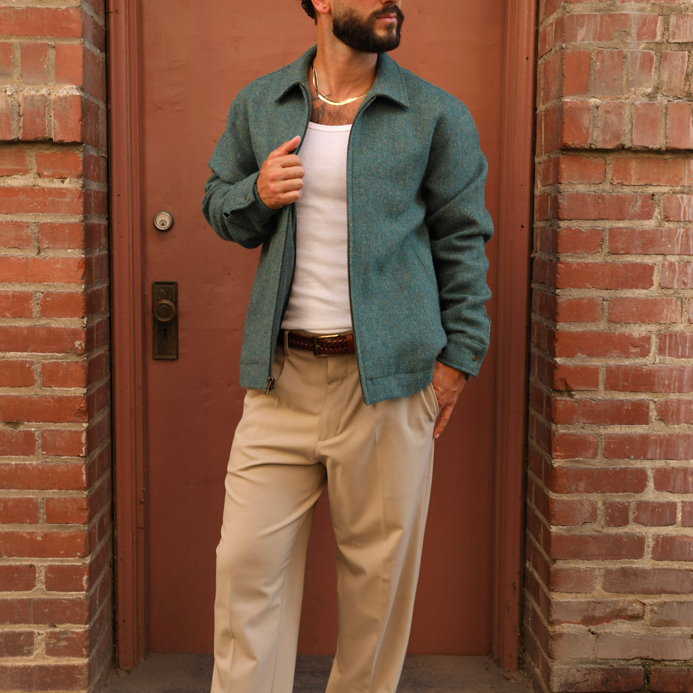 A man wearing a dark green overshirt, beige trousers, a white tank top, a black baseball cap, sunglasses, and brown loafers stands in front of a brick building with a brown door.