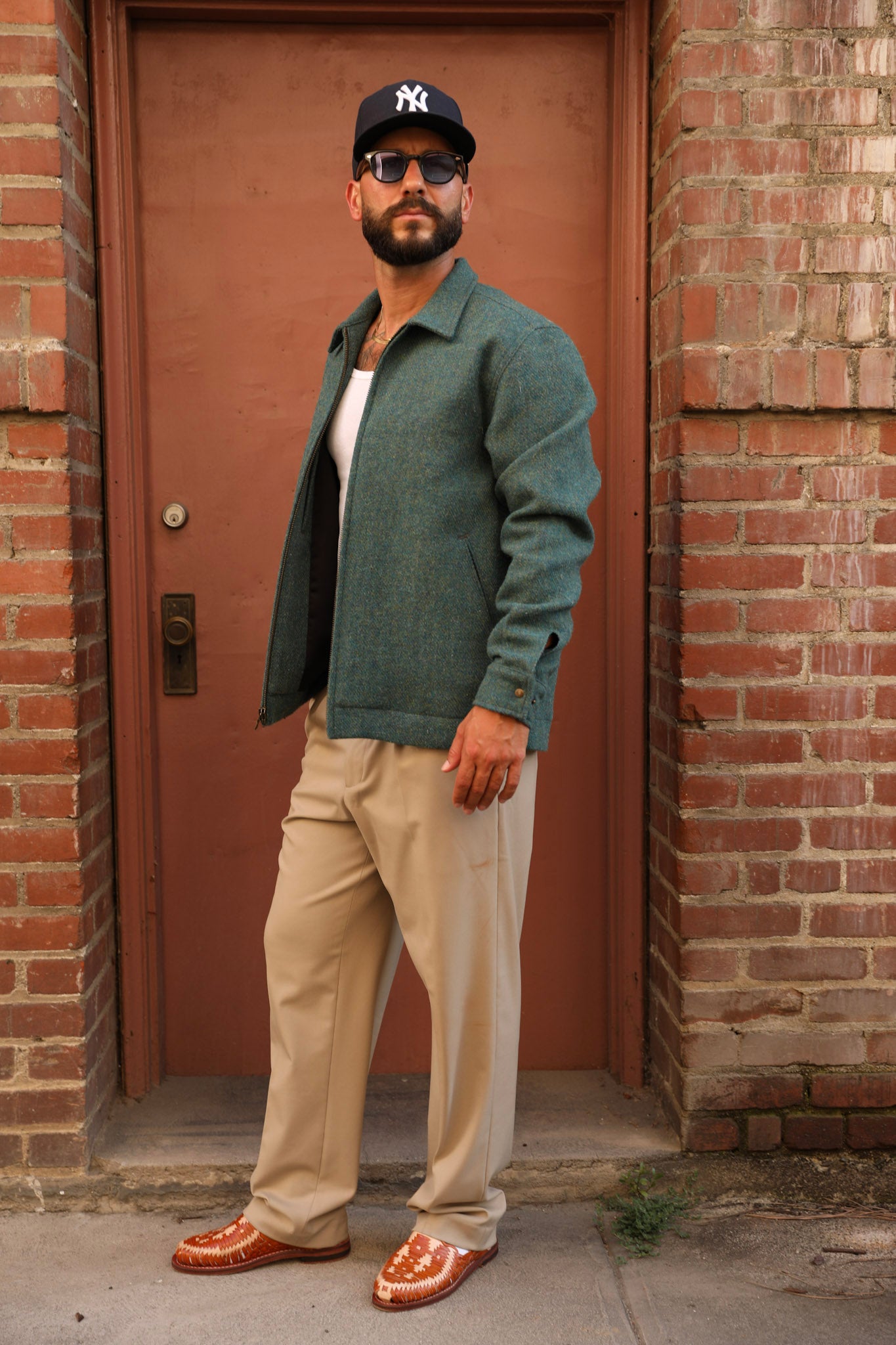 A man wearing a dark green textured overshirt, beige trousers, brown leather loafers, and a black New York Yankees baseball cap stands in front of a red-brown door framed by brick walls.