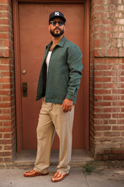 A man wearing a dark green textured overshirt, beige trousers, brown leather loafers, and a black New York Yankees baseball cap stands in front of a red-brown door framed by brick walls.